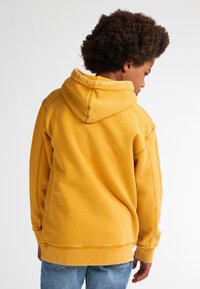 Felpa gialla con una texture morbida, dotata di cappuccio e polsini a costine. La parte posteriore presenta cuciture visibili e una vestibilità comoda. Sotto sono indossati jeans in denim.