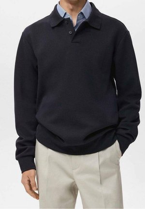 Muž v černém polokošili s dlouhým rukávem přes modrou proužkovanou košili s límečkem a béžových kalhotách, stojící s jednou rukou v kapse.