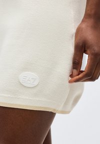 Κοντινό πλάνο του χεριού ενός ατόμου που ξεκουράζεται κοντά στο τελείωμα ενός υφασμάτινου σορτς σε ελαφρώς εκρού απόχρωση με υφή, με ένα λογότυπο της EA7 Emporio Armani.