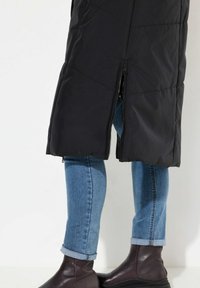 Svart vadderad kappa med diagonala sömmar, sidodragkedja och hög slits. Buren över blå jeans med rullade ärmar och mörkbruna ankelstövlar.