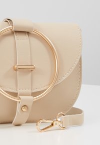 Beige läder crossbody-väska med rund guldfärgad beslag, klaffstängning och justerbar axelrem med klipsfäste.