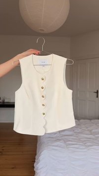 Krēmveida bezroku blūze ar viļņotu priekšējo malu un piecām dekoratīvām zelta pogām, pakārta uz pakaramā telpās virs gultas.