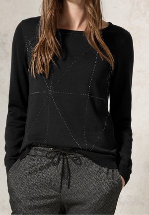 Mujer con top de manga larga negro con patrón geométrico de puntos y pantalones grises de espiga con cordón, manos en los bolsillos, sobre fondo claro.