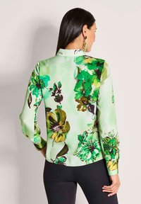 Vrouw met lang donker haar, gekleed in een groene bloemenblouse met lange mouwen en een zwarte broek, vanachter weergegeven met zichtbare oorbellen.