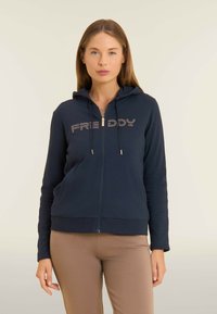 Donna che indossa una felpa con zip blu navy con "FREDDY" in strass e pantaloni beige, in piedi contro uno sfondo chiaro e semplice.