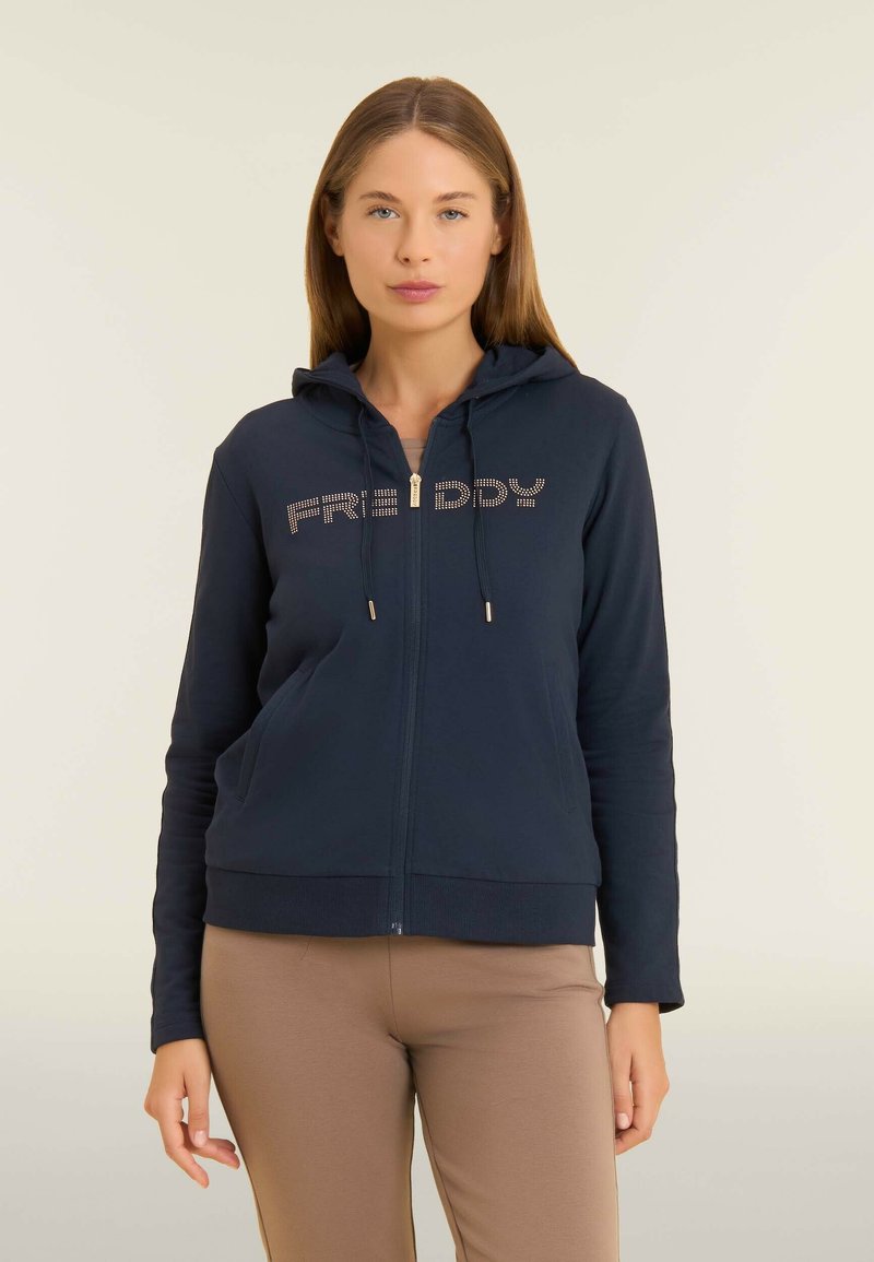 Donna che indossa una felpa con zip blu navy con "FREDDY" in strass e pantaloni beige, in piedi contro uno sfondo chiaro e semplice.