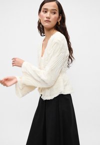 Biela, textúrovaná blúzka s výstrihom do V a zvonovými rukávmi, s vlnitým, plisovaným dizajnom. Nosená cez čiernu sukňu.