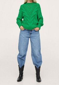 Groen gebreid sweater met textuurpatronen en geribde manchetten, gecombineerd met lichtblauwe wijde jeans en zwarte enkellaarzen.