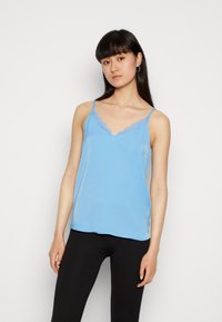 Camisola de tom azul claro com alças finas, apresentando um decote em V e detalhes em renda. Tecido suave com uma textura lisa. Usada com leggings pretas.