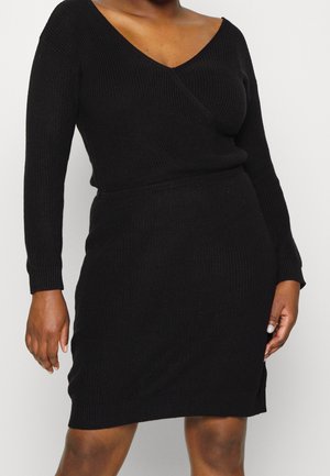 Robe en maille noire à manches longues avec encolure en V portée par une personne, montrant le torse et le haut des jambes sur un fond clair uni.