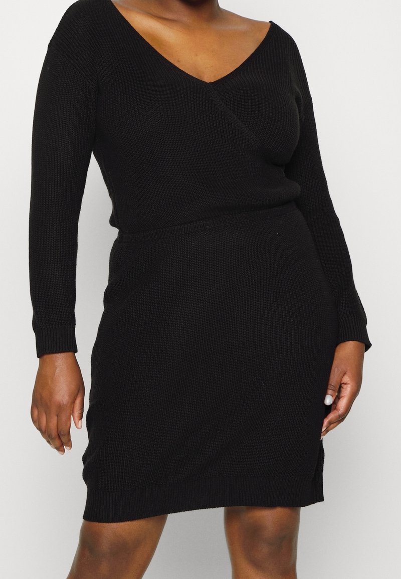 Robe en maille noire à manches longues avec encolure en V portée par une personne, montrant le torse et le haut des jambes sur un fond clair uni.