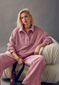 Chemise oversize en velours côtelé rose avec col, associée à un pantalon large assorti. Assise sur une chaise texturée couleur crème, tenant un sac.