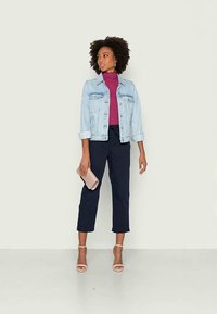 Hellblaues Jeansjackett, Pflaumenfarbener Rollkragenpullover, marineblaue Cropped-Hosen und beiger Clutch. Sandalen mit offenen Zehen und lockige Haare runden den Look ab.