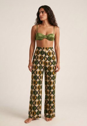 Mujer con cabello largo y rizado lleva una camiseta corta de satén verde y pantalones de pierna ancha de talle alto con patrones de círculos amarillos y verdes, de pie descalza.