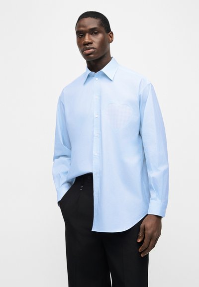 Άνδρας με γαλάζιο μακρυμάνικο πουκάμισο με διακριτικό περίγραμμα καρδιάς στο στήθος και μαύρο παντελόνι, όρθιος με το χέρι στην τσέπη.