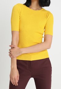 Ribbed, short-sleeve top in bright yellow cotton. Features a round neckline and a fitted silhouette, paired with dark burgundy pants.