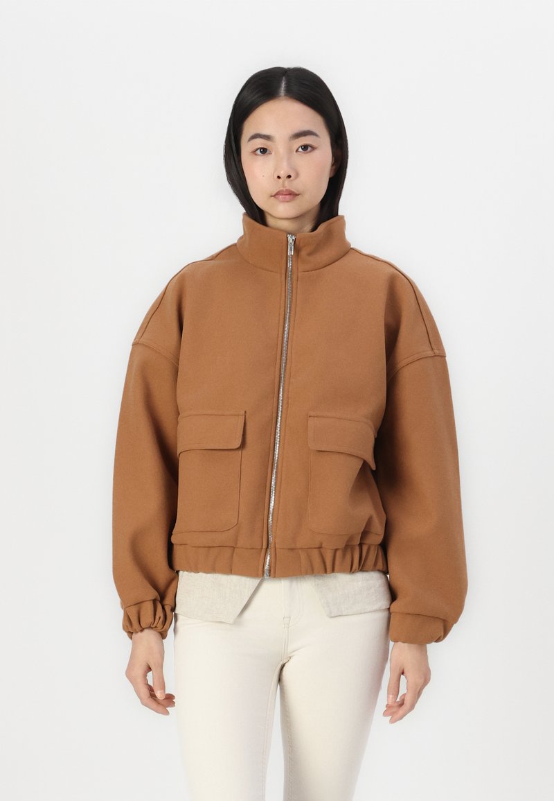 Veste brune zippée avec un col haut, deux poches avant, un ourlet élastiqué et des manches surdimensionnées, fabriquée en tissu doux.