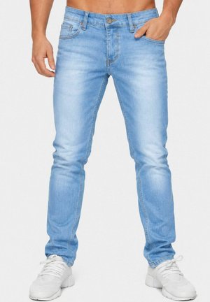 Man draagt lichtblauwe slim-fit jeans en witte sneakers met één hand in de zak tegen een effen achtergrond.