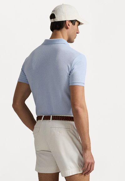 Un tricou polo cu mânecă scurtă de culoare albastră deschisă, asociat cu pantaloni scurți bej deschis și o curea împletită maro, purtat cu o șapcă albă. Textura materialului este netedă.
