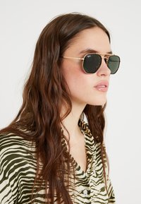 Sunglasses with black lenses and gold metal frames, featuring a geometric shape. Worn by a person in a zebra-striped shirt with green tones.