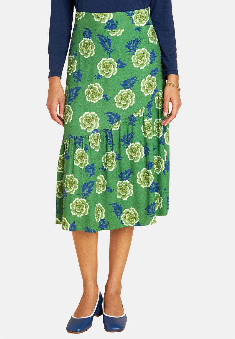 Eine Person trägt einen grünen Blumenkleid mit weißen und blauen Blumenmustern, kombiniert mit einem marineblauen Langarmoberteil und blauen Schuhen.