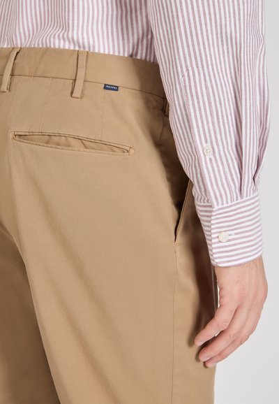 Uomo che indossa pantaloni beige con tasca posteriore a filetto e manica di camicia a righe rosse e bianche con bottoni visibile.
