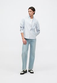 Helles blaues Hemd mit langen Ärmeln, über einem weißen T-Shirt getragen. Kombiniert mit hellblauen geraden Jeans und schwarzen Schuhen mit weißen Akzenten.