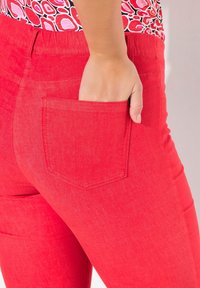 Röda denimbyxor med en figurnära design, utrustade med en synlig bakficka och kontrasterande sömmar. Tygets textur ser slät och något strukturerad ut.