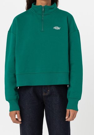 Personne portant un sweat-shirt à col zippé court bleu sarcelle avec un petit logo blanc sur la poitrine et un jean bleu foncé.