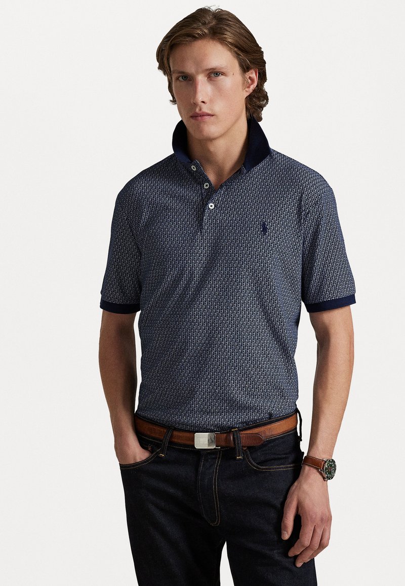 Polo de padrão marinho com colarinho contrastante, mangas curtas e uma pala com três botões, combinado com jeans escuros e um cinto castanho.