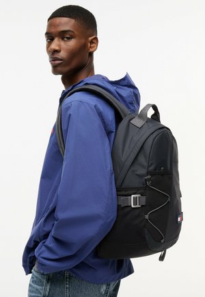 Mężczyzna w niebieskiej kurtce i dżinsach niesie na plecach czarny plecak z siateczkowymi kieszeniami i paskami z klamrami.