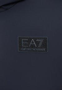Tecido azul-marinho com um retângulo preto em relevo contendo as letras "EA7 EMPORIO ARMANI" e um logótipo, superfície texturizada com detalhes finos.