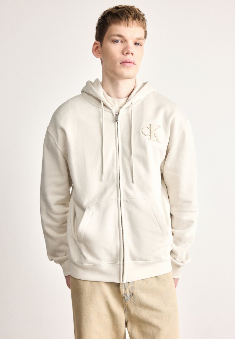 Világosbézs cipzáros kapucnis pulóver puha anyagból, állítható zsinórokkal és logóval a bal mellrészen. Két első zsebbel és bordázott szegéllyel rendelkezik.