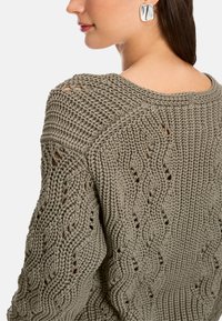 Gewobener Pullover in Olivgrün mit einem strukturierten, offenen Muster und einem gerippten Kragen. Verfügt über leicht fallende Schultern und feine Details.