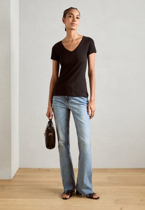 Tricou negru cu mânecă scurtă și decolteu în V, blugi evazați de culoare albastră estompată, geantă maro cu mâner și sandale negre fără toc pe un podea din lemn.