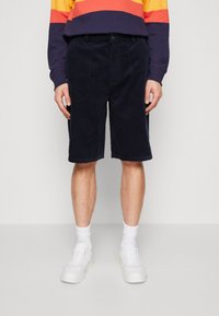 Pantalones cortos de corduroy azul marino con corte recto, que cuentan con bolsillos frontales y una textura sólida. Estilizados con zapatillas blancas y calcetines hasta el tobillo.