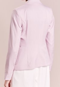 Světle růžový blazer s přiléhavým střihem, dlouhými rukávy a jednou kapsou. Hladká textura a minimální detaily na zadní straně.