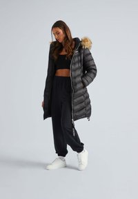 Zwarte pufferjas met een met bont gevoerde capuchon, ritssluiting en elastische manchet. Draagt over een zwarte crop top en gecombineerd met zwarte sweatpants en witte sneakers.