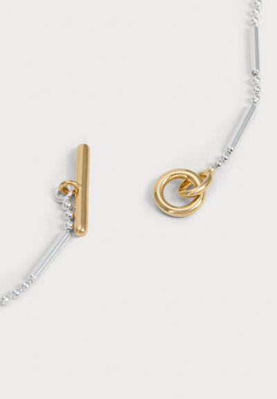 Collier bicolore en argent et en or, avec une chaîne boule, des barres allongées et des pendentifs circulaires entrelacés sur un fond clair.