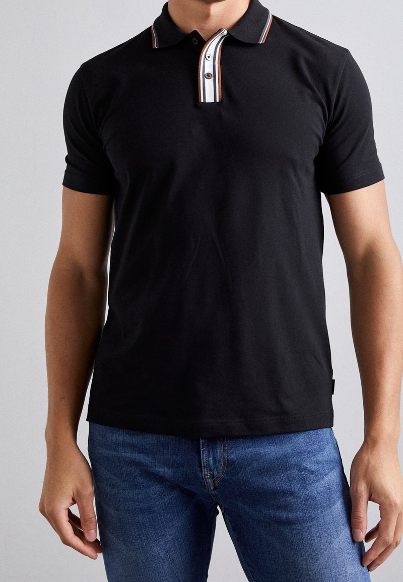 Homme portant un polo noir à manches courtes avec un col rayé et un jean bleu, debout devant un fond uni.