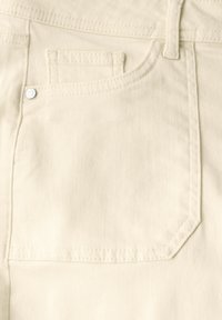 La tela de denimas beige presenta un bolsillo trasero con un acento de botón plateado, una textura suave y costuras limpias a lo largo de los bordes.