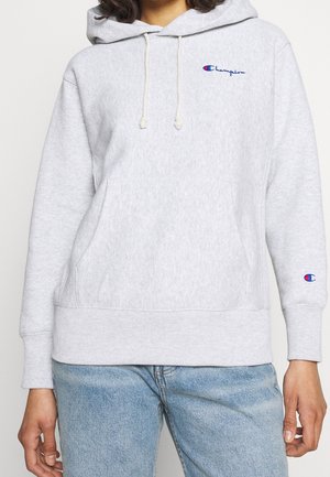 Person iført en lysegrå Champion hættetrøje med frontlomme og blåt logo, kombineret med lyseblå jeans.