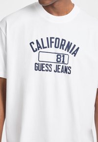 Bílý bavlněný tričko s krátkými rukávy, se námořnickým grafickým potiskem, který zahrnuje "CALIFORNIA," "81," a "GUESS JEANS."