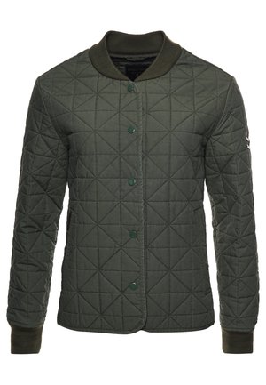 Personne portant une veste matelassée olive, un pantalon court foncé, des bottines noires et un pull foncé, debout devant un panneau en bois.