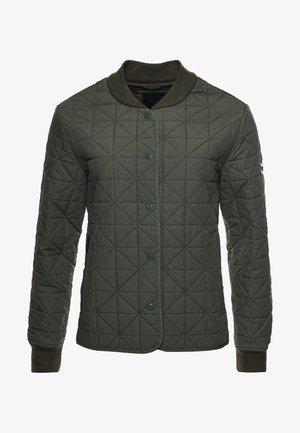 Person iført en olivengrøn quiltet jakke, mørke korte bukser, sorte ankelstøvler og en mørk sweater, stående mod en baggrund af træpaneler.