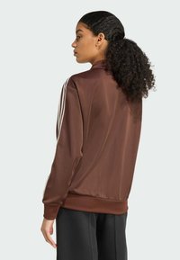 Veste de sport en satin marron avec trois bandes blanches sur les manches, poignets côtelés et coupe décontractée, associée à un pantalon noir.