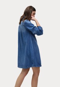 Sukienka jeansowa o luźnym kroju, z długimi rękawami i kołnierzykiem zapinanym na guziki. Materiał jest w średnim niebieskim kolorze, z lekko wyblakłym wyglądem i subtelnym przeszyciem.
