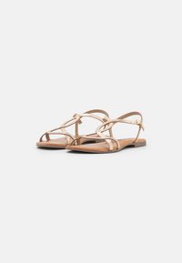 Sandales plates en métal doré avec plusieurs bandes croisées, une sangle de cheville à boucle, et une semelle en cuir naturel brun clair.