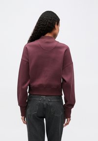 Mujer con pelo rizado largo de espaldas, vistiendo un sudadero morado oscuro y unos vaqueros negros de cintura alta, contra un fondo liso.