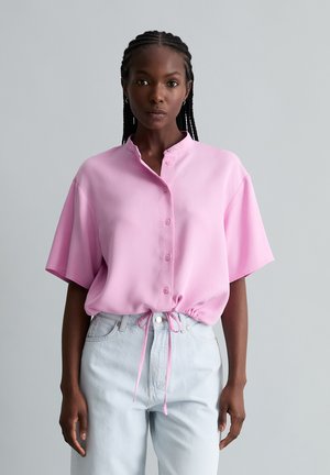 Frau mit geflochtenem Haar, die eine hellrosa kurzärmelige, geknöpfte Bluse trägt, die an der Taille gebunden ist, und hellblaue, hoch taillierte Jeans.